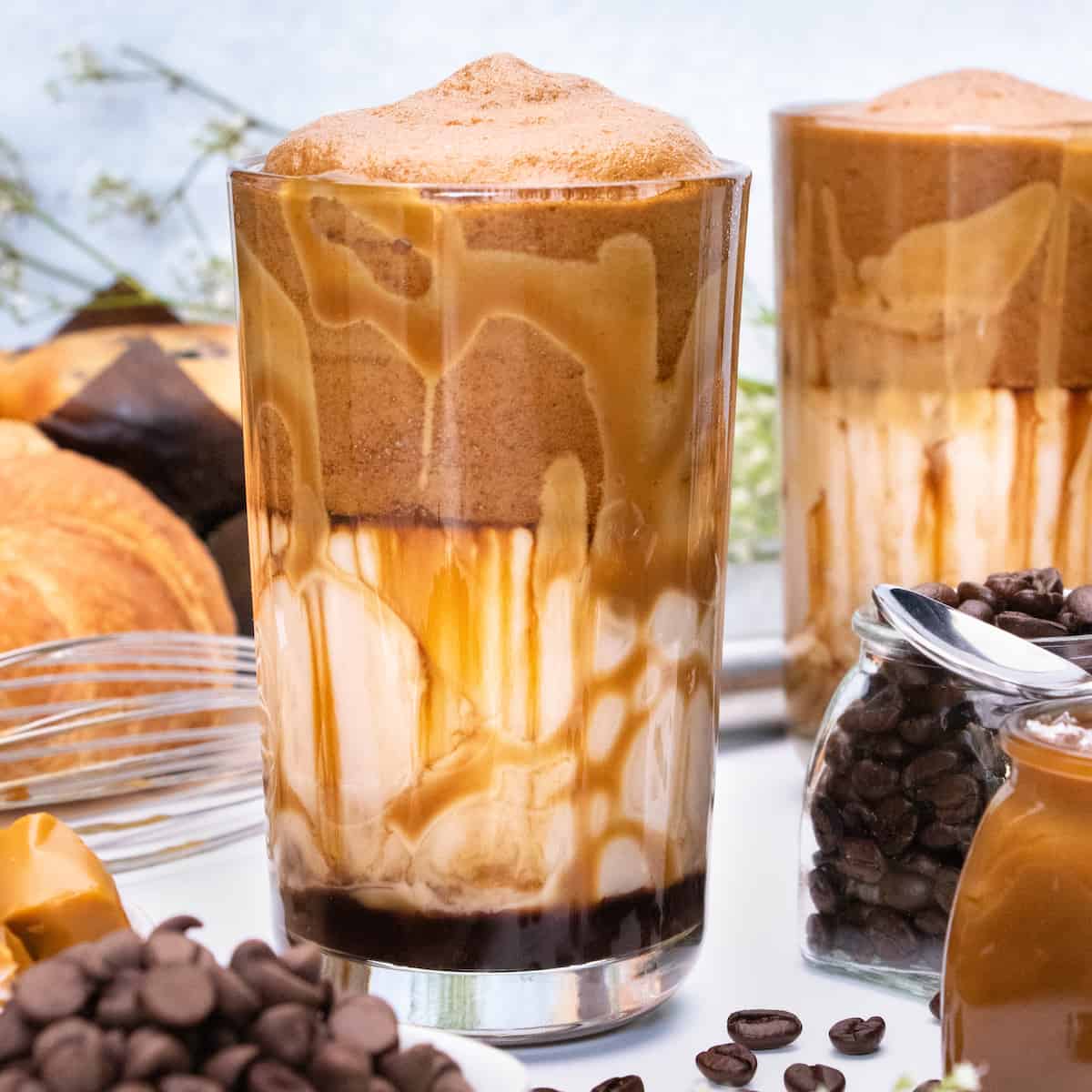 Two whipped salted caramel mocha lattes. They are made with chocolate syrup, salted caramel sauce, milk, and whipped coffee. Ingredients are in the foreground.